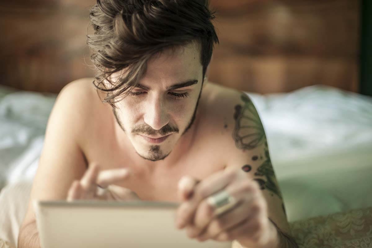 Ein junger Mann mit Tattoos liegt im Bett und liest auf seinem Tablet.