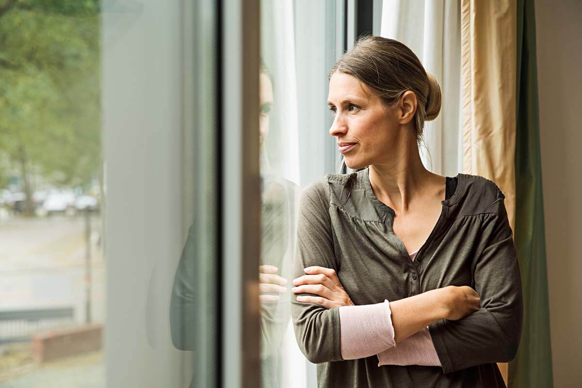 Eine Frau steht mit verschränkten Armen am Fenster und schaut hinaus.