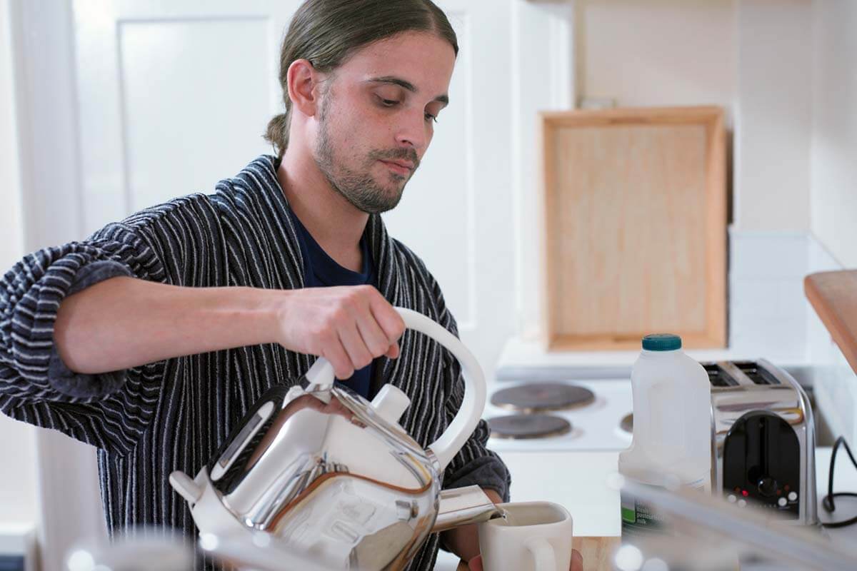 Ein Mann im Bademantel gießt sich heißes Wasser in eine Tasse.