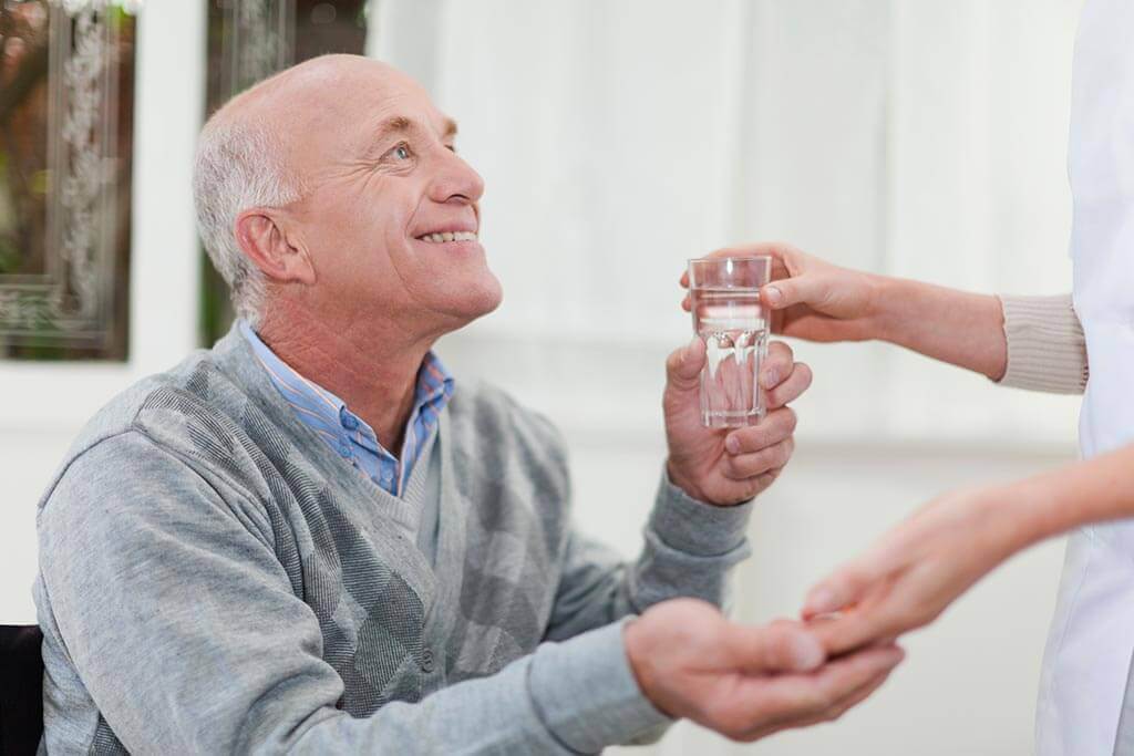 Eine Frau reicht einem älteren Mann ein Glas Wasser und Tabletten