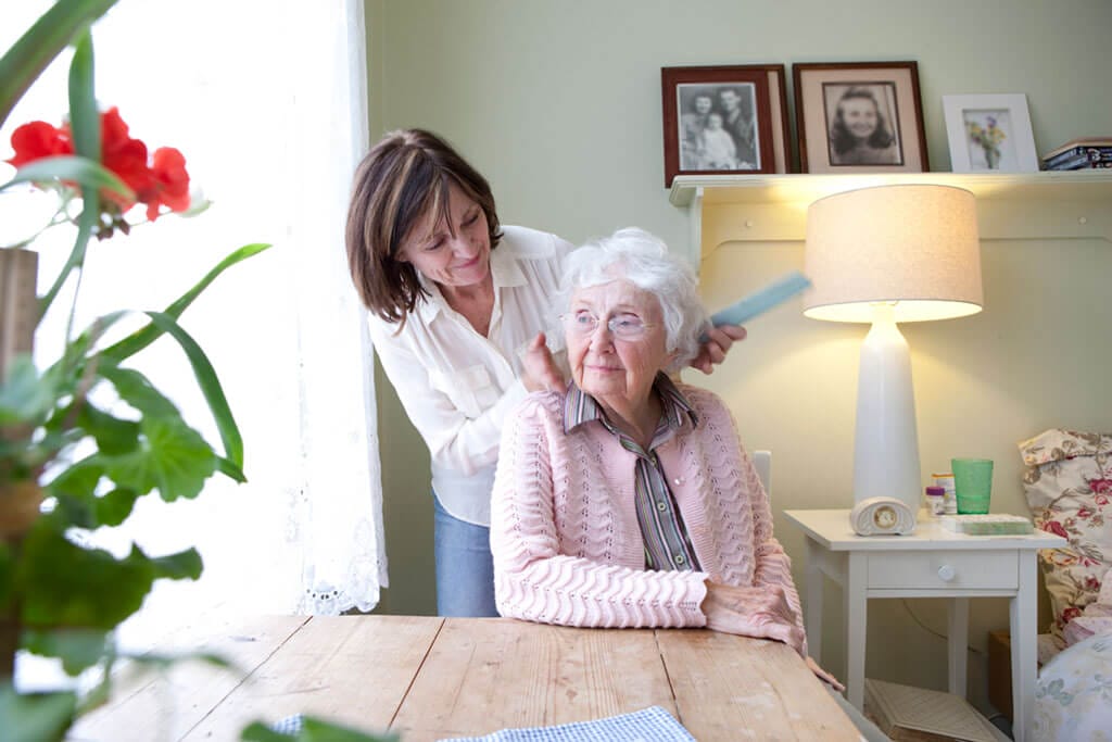 Eine Seniorin sitzt am Tisch. während ihre Tochter ihr über die Wange streicht